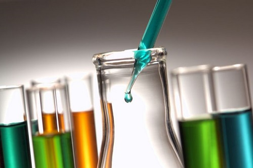 Several chemical test tubes contain different colored liquids, and a dropper is dropping liquid into a container